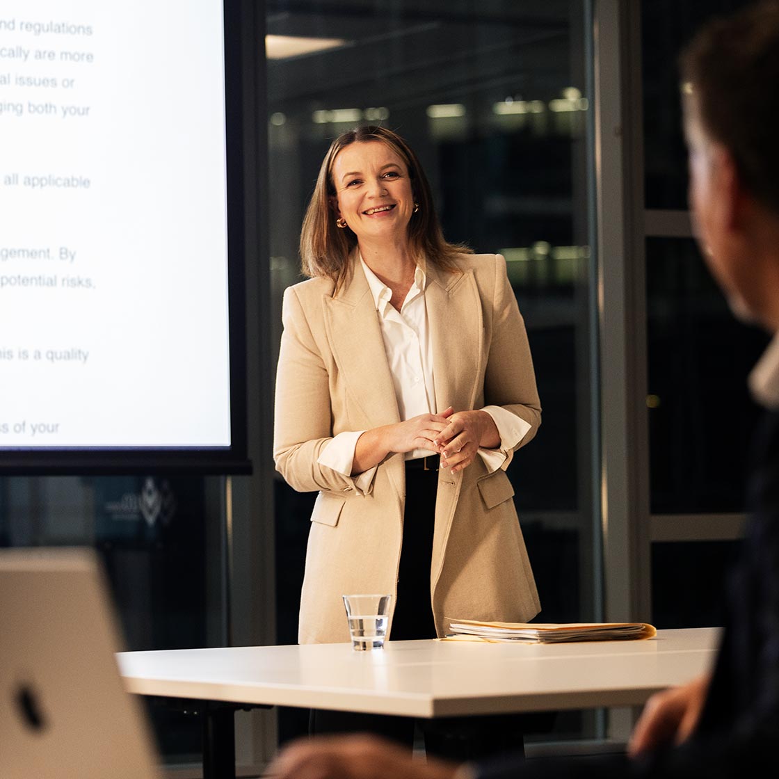 A middle-aged woman stands in front of a projector screen giving a lecture.
